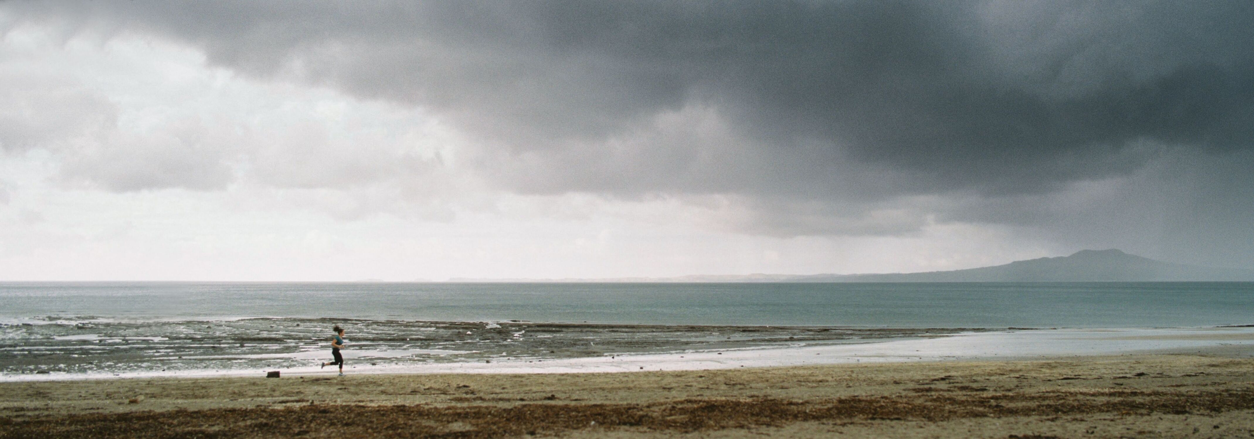 Panorama of a woman running up the beach with a cloudy mountain in the background