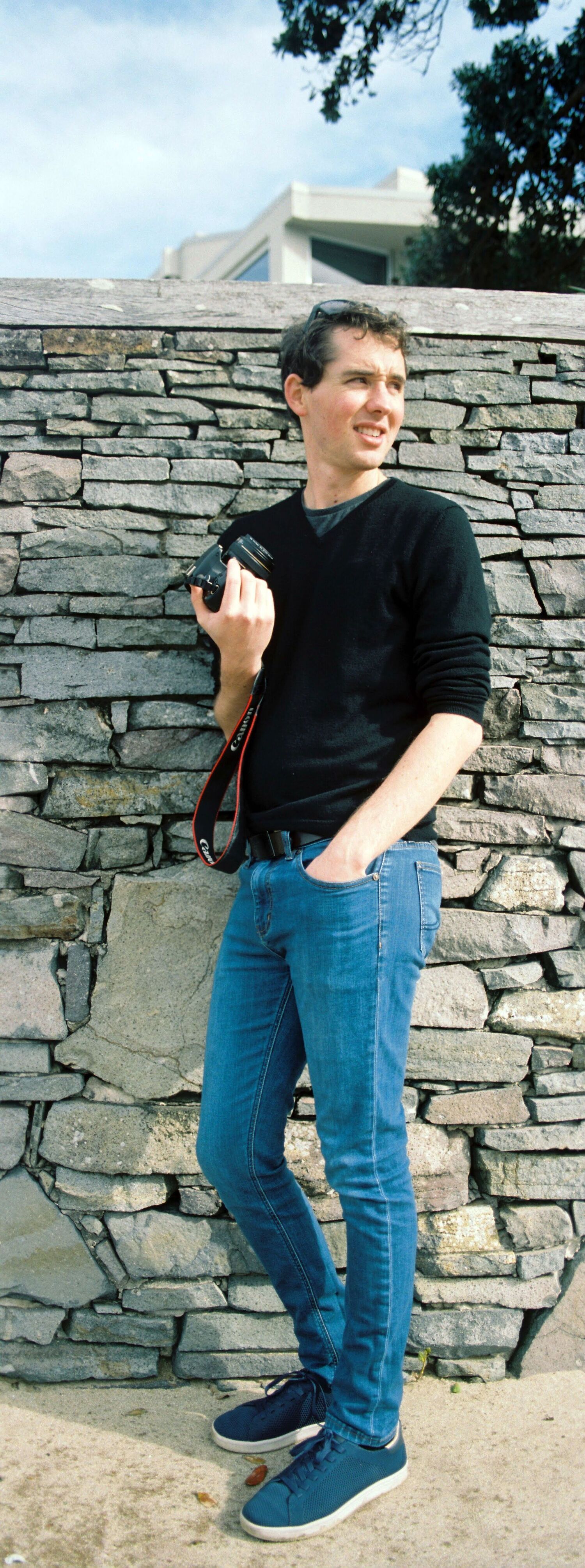 Vertical panorama of Byron posing with his camera up against a stone wall